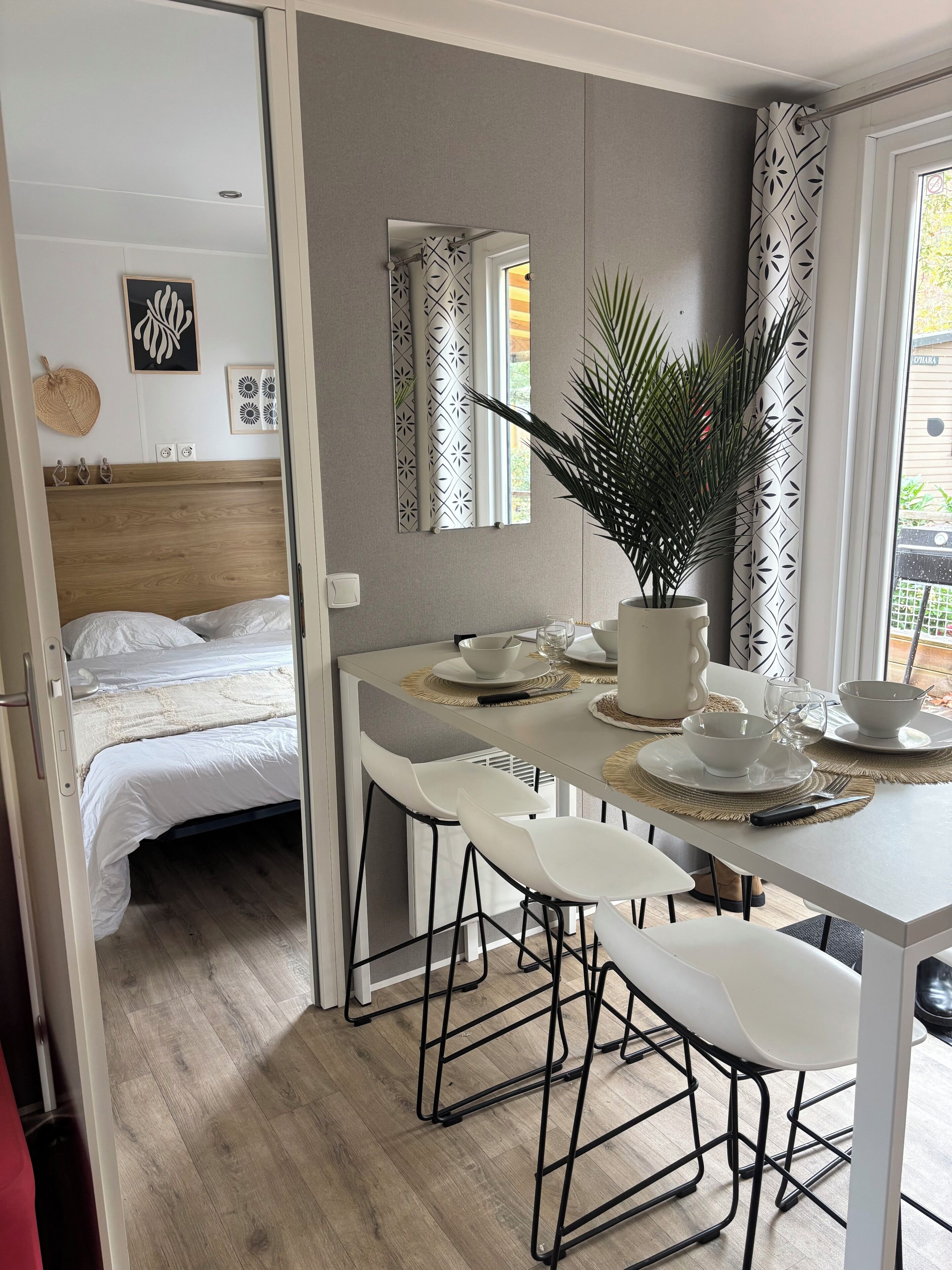 Una habitación moderna con una cama, una mesa de comedor con sillas, una planta en maceta y un ventanal con cortinas.