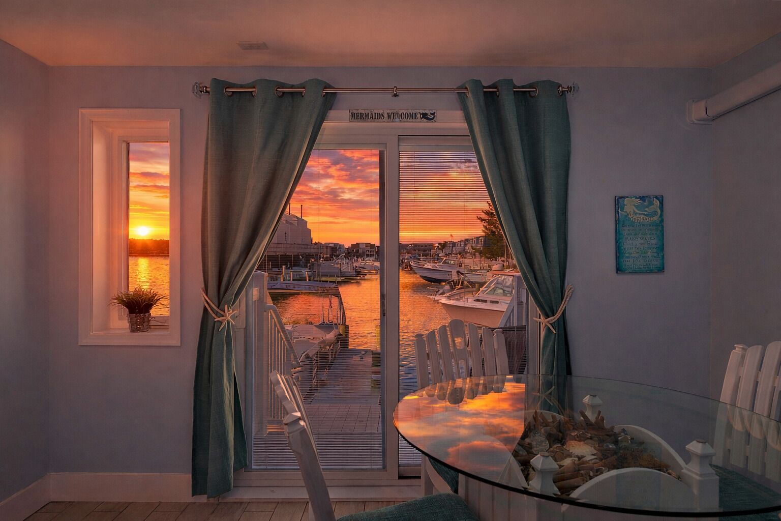 A room with a view of a marina at sunset, a glass table, and a sign reading 'Mermaids Welcome'.