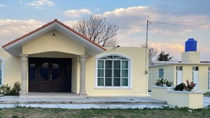 A two-story house with a beige facade, a central entrance with a dark door, and a red roof. - Beautiful, modern 2-bedroom home located on a private street. (Teoloyucan)