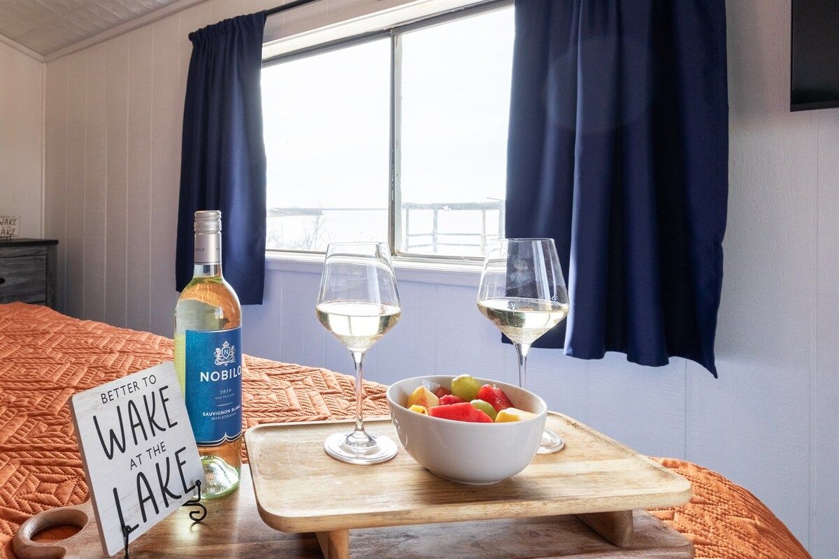 A bed with a sign that reads 'Better to wake at the lake,' a bottle of wine, two glasses, and a bowl of fruit on a wooden tray.