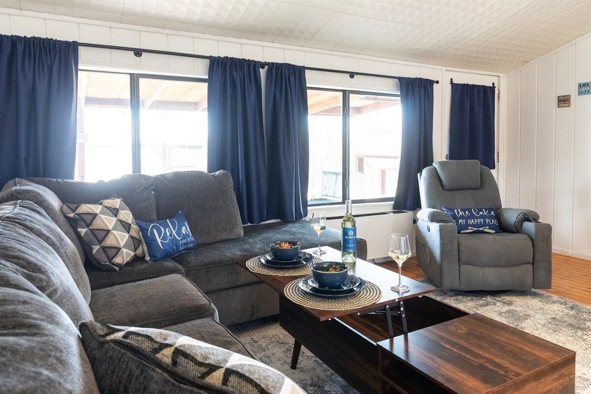 Ein Wohnzimmer mit einer Couch, einem Sessel und einem Couchtisch. Auf dem Tisch stehen zwei Schalen mit Futter und eine Weinflasche.