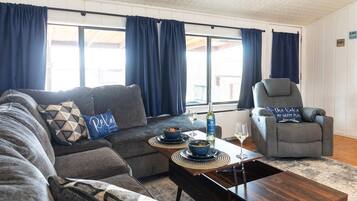 Ein Wohnzimmer mit einer Couch, einem Sessel und einem Couchtisch. Auf dem Tisch stehen zwei Schalen mit Futter und eine Weinflasche.