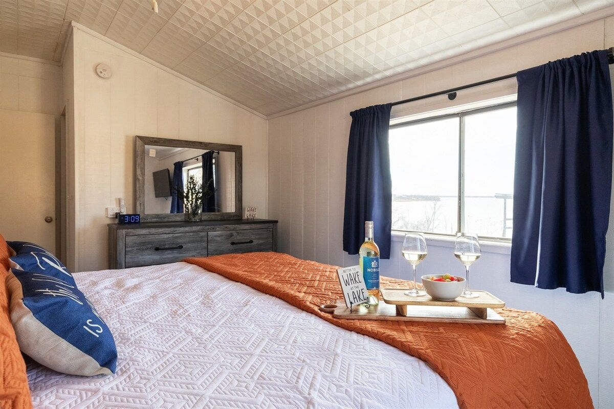 Et hotelværelse med seng, kommode, spejl, et vindue med gardiner og et bakke med en flaske og glas.