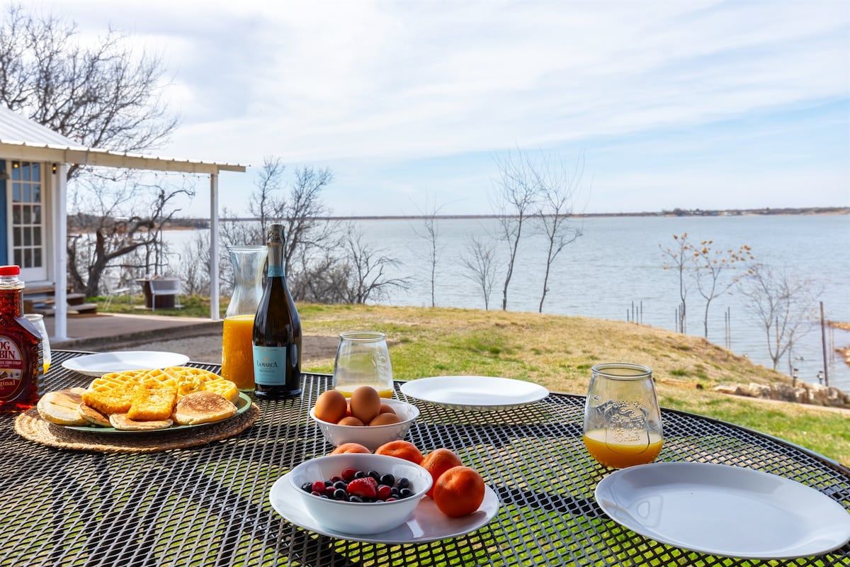 Un petit-déjeuner composé de gaufres, d’œufs, de fruits rouges et de jus, servi sur une table près d’un lac.