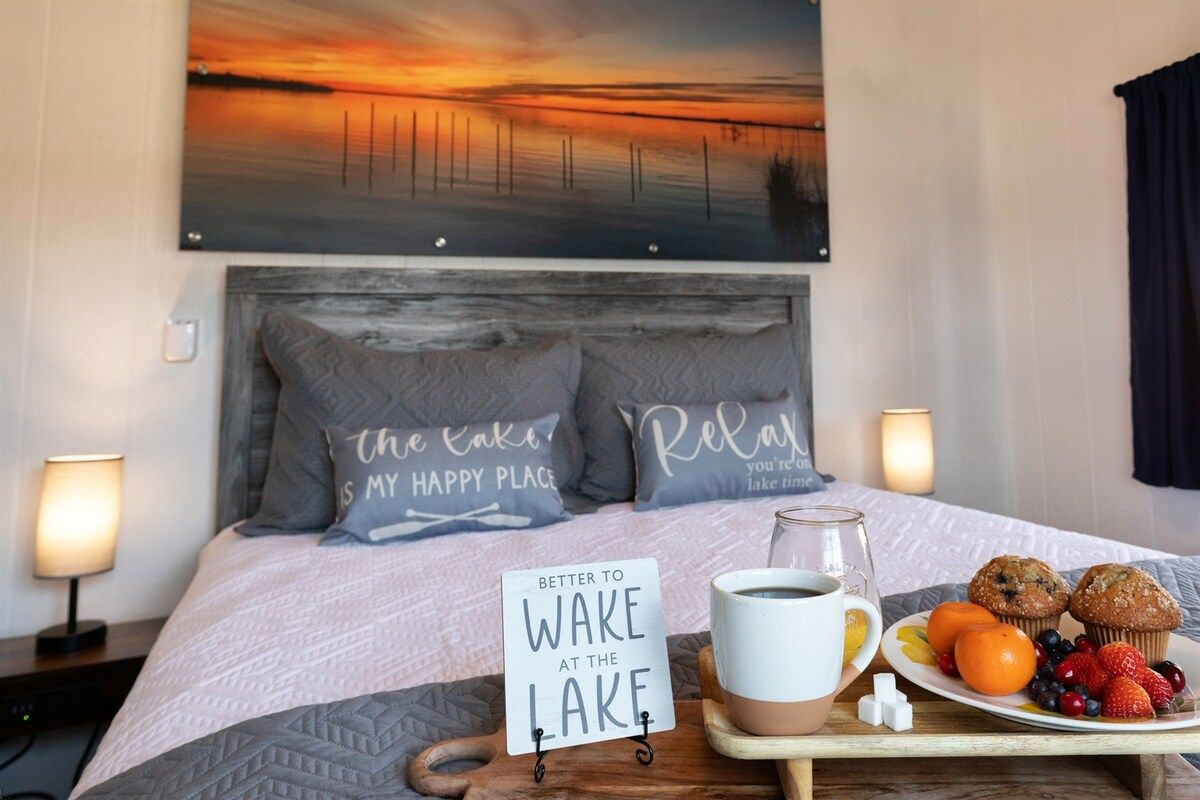 A hotel room with a bed, a painting of a sunset, pillows with text, a tray with muffins and fruit, and a sign on the bed.