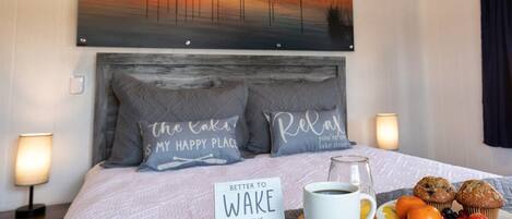 Habitación de hotel con una cama, un cuadro de un atardecer, almohadas con texto, una bandeja con muffins y fruta, y un cartel sobre la cama.
