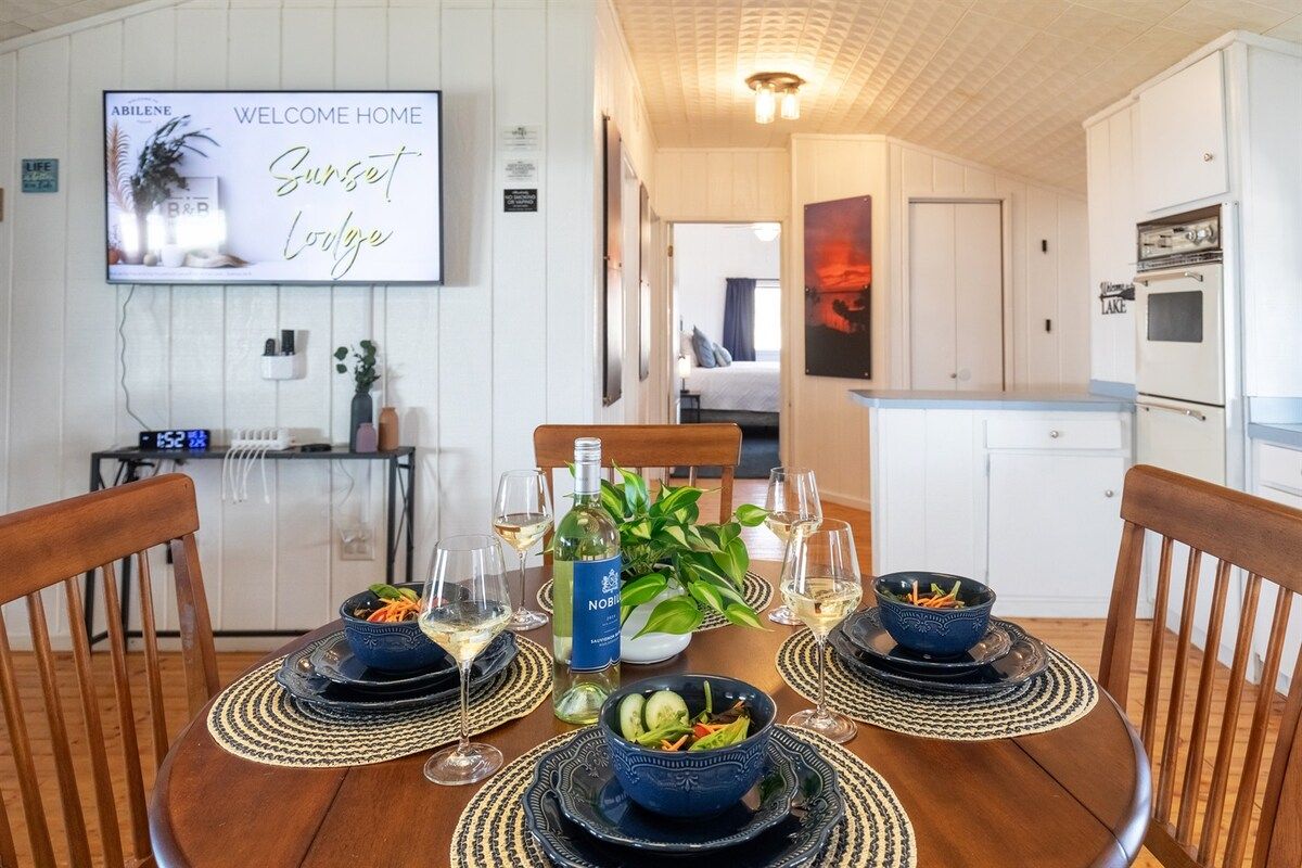 A dining area with a table set for four, a television displaying 'Welcome Home Sunset Lodge,' and a kitchen in the background.