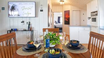 A dining area with a table set for four, a television displaying 'Welcome Home Sunset Lodge,' and a kitchen in the background.