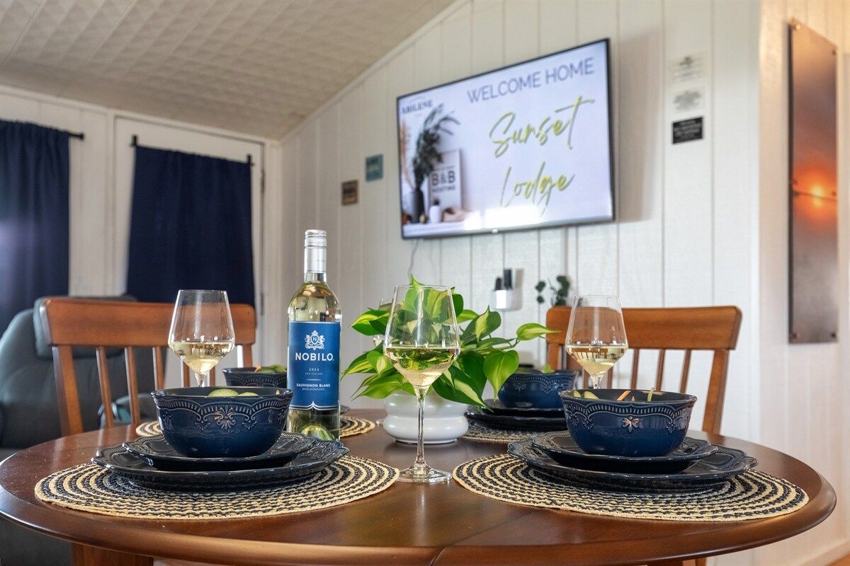 Une table à manger dressée avec des bols et des assiettes bleus, une bouteille de vin, un panneau indiquant « Bienvenue à la maison » et une plante en arrière-plan.