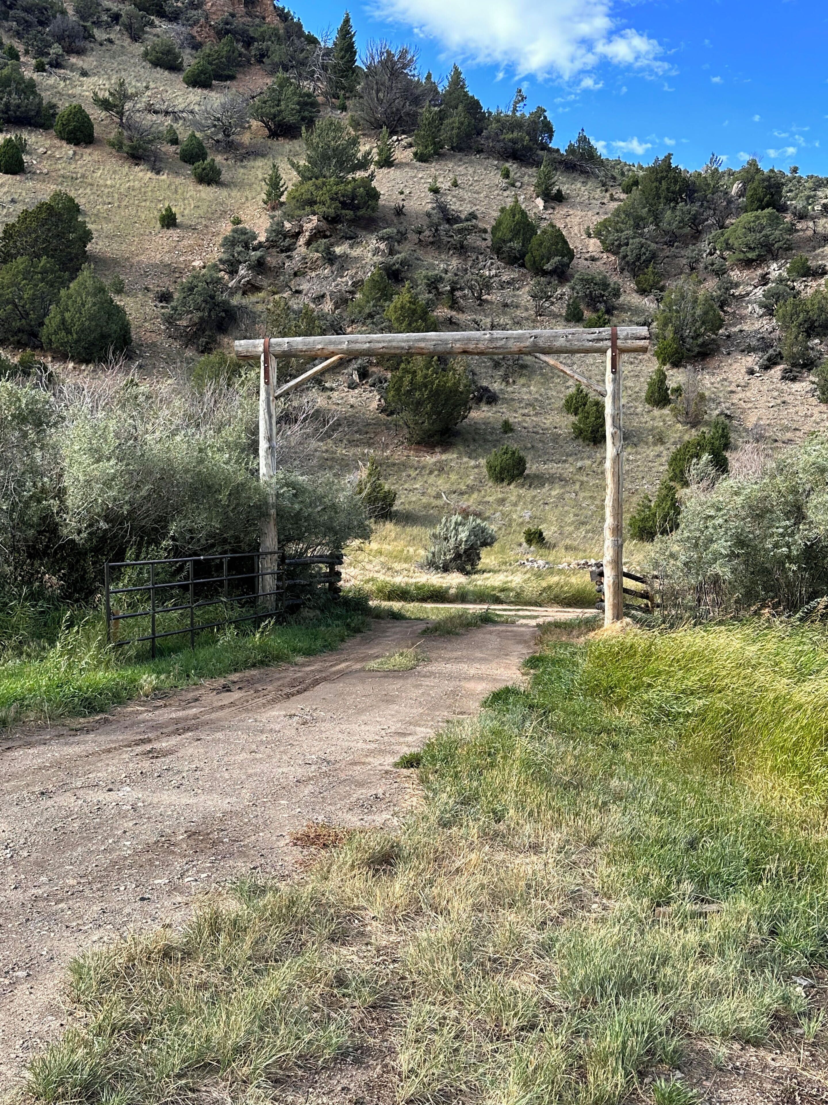 A dirt path leading to a wooden gate in a natural setting with sparse vegetation and a hill in the background.