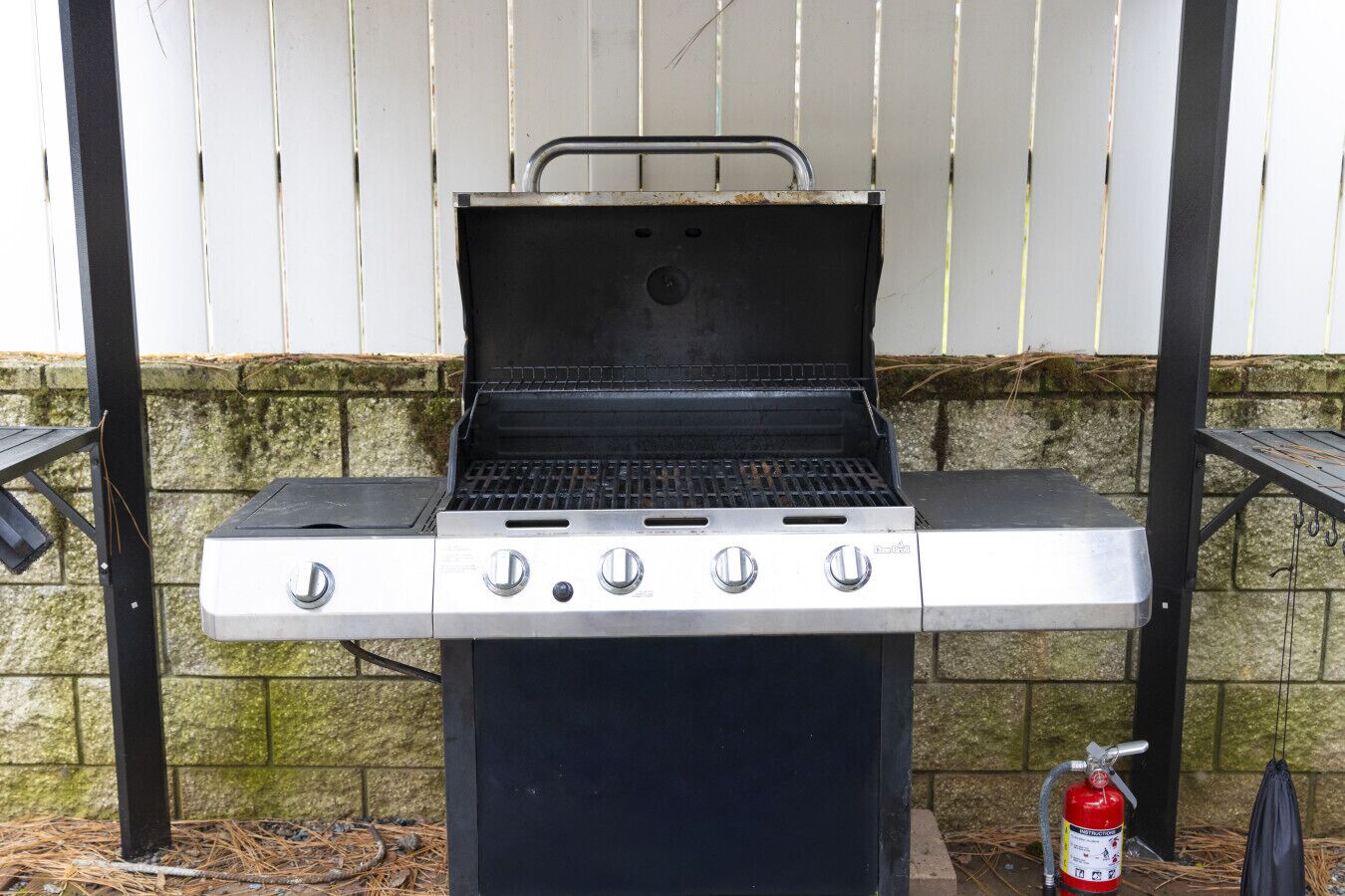A barbecue grill with four burners, a side shelf, and a fire extinguisher nearby.