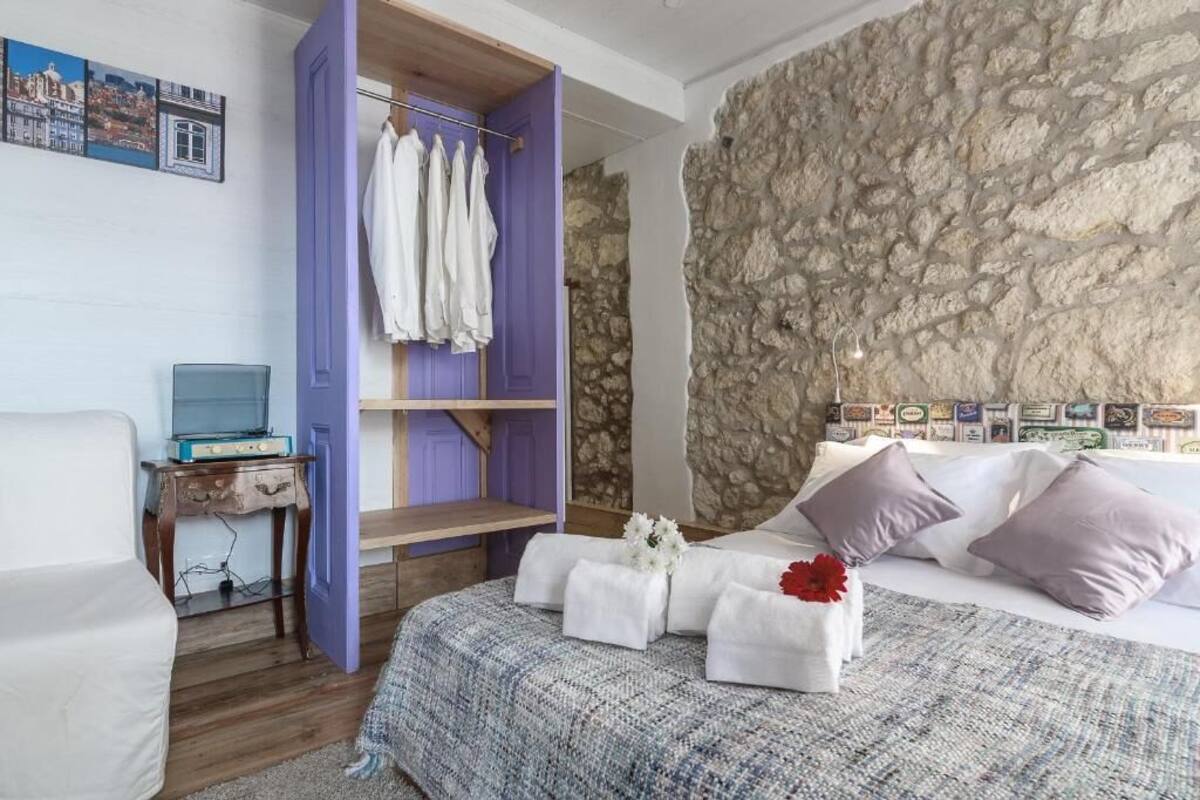 A bedroom with a stone wall, a bed with white and purple bedding, a wooden nightstand, and a purple wardrobe.