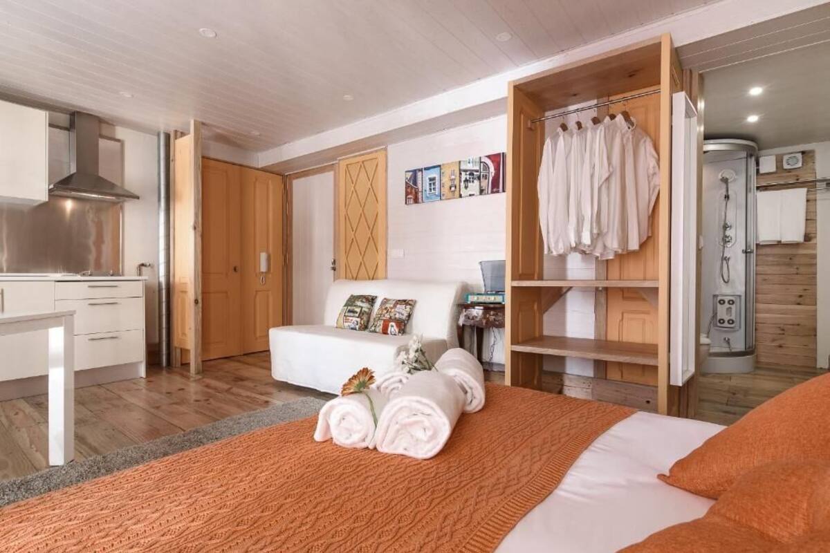 A modern hotel room with a bed, a sofa, a wooden wardrobe, a shower area, and a kitchenette.