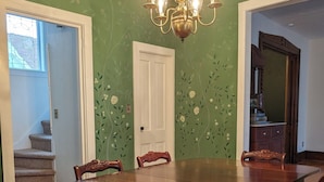 A dining room with a wooden table and chairs, a chandelier, and a green floral wall. - The Ainsworth House - Historic Victorian 3BR Gem (Stoughton)