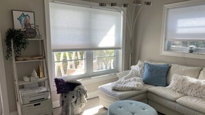 Ein modernes Wohnzimmer mit einem weißen Sofa, einem blauen Kissen, einem grauen Hocker und einem weißen Bücherregal, sowie einem Fenster mit Jalousien.