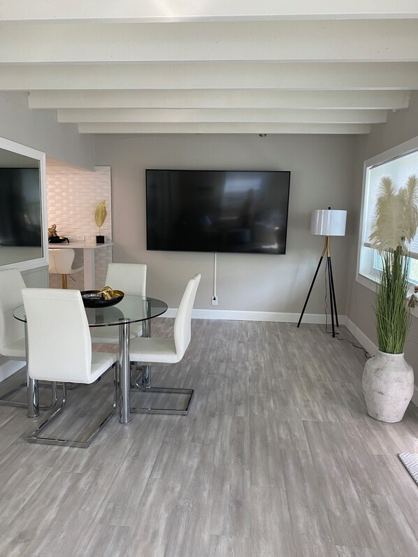 A modern living room with a glass dining table, white chairs, a flat-screen TV, a floor lamp, and a potted plant.