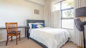 Een slaapkamer met een bed, een bureau, een stoel, een ventilatorkachel en een raam met gordijnen.