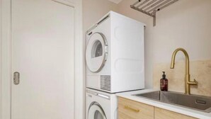 Een kleine wasruimte met gestapelde wasmachines, een wastafel met een gouden kraan, en houten kastjes.