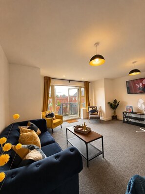 Ein Wohnzimmer mit einer Couch, einem Couchtisch und einem Sessel. Es gibt einen Balkon mit Blick auf Gebäude.