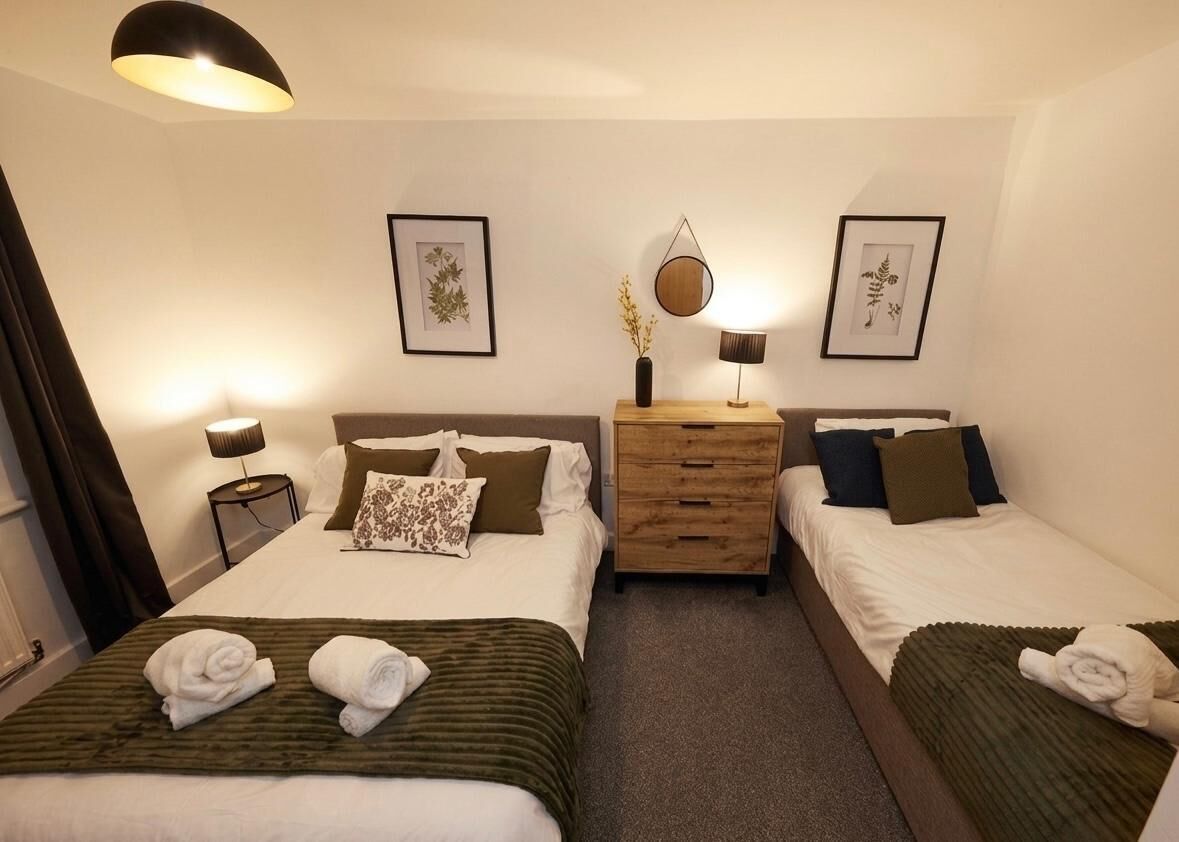 Hotellihuone, jossa on kaksi sänkyä, puinen lipasto, tauluja seinillä ja lamppu huoneen molemmilla puolilla.