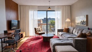 Moderni hotellihuone, jossa on suuri ikkuna, sohva, nojatuoli, pieni pöytä ja televisio.