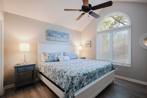 A bedroom with a bed, two bedside lamps, a ceiling fan, a window with blinds, and a painting on the wall. - Sea Pines Serenity w/pool access & beach shuttle (Hilton Head Island)
