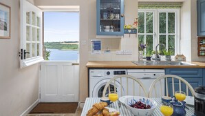 A kitchen with a view of a lake, a washing machine, a sink, and breakfast items on the table. - Charming Riverside Cottage in the Heart of Pembrokeshire (Cosheston)