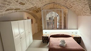 Een slaapkamer met een stenen plafond, een bed met een bruine plaid, een witte kledingkast en een klein nachtkastje met een lamp.