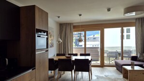 Eine moderne Küche mit Holzschränken, ein Essbereich mit Tisch und Stühlen, ein Sofa und eine große Schiebeglasdoor zu einem Balkon.