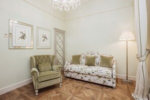 Een kamer met een groene fauteuil, een bloemenbank, een kroonluchter, bloemistisch kunstwerk in een lijst en een vloerlamp.