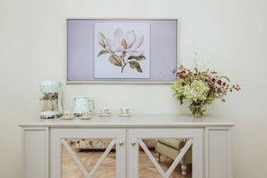 A white console table with a coffee maker, cups, and saucers, a framed floral artwork above, and a vase with flowers to the right. - Deluxe Junior Suite with Terrace & Balcony in Sorrento Center (Sorrento)