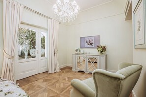 A room with a wooden parquet floor, a white cabinet with a glass top, a green armchair, a chandelier, and a window with curtains. - Deluxe Junior Suite with Terrace & Balcony in Sorrento Center (Sorrento)