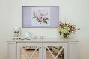 Een witte console tafel met een koffiezetapparaat, kopjes en schoteltjes, een lijst met bloemkunstwerk erboven, en een bloemenvaas aan de rechterkant.
