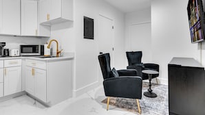 Una cocina moderna con armarios blancos y tiradores dorados, una silla negra con una mesa redonda, y un televisor de pantalla plana montado en la pared.
