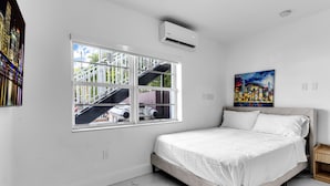 Un dormitorio con cama, ventana con vista a edificios, aire acondicionado y un cuadro en la pared.