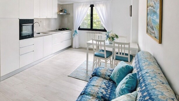 Uma cozinha moderna com armários brancos, uma área de jantar com mesa e cadeiras brancas, e um sofá azul com padrão.