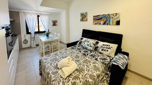 Quarto com cama, cabeceira, janela com cortinas, mesa de jantar com cadeiras e área de cozinha.