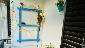 Uma pequena varanda com mesa e cadeiras brancas, uma prateleira azul com luzes decorativas e um vaso com planta.