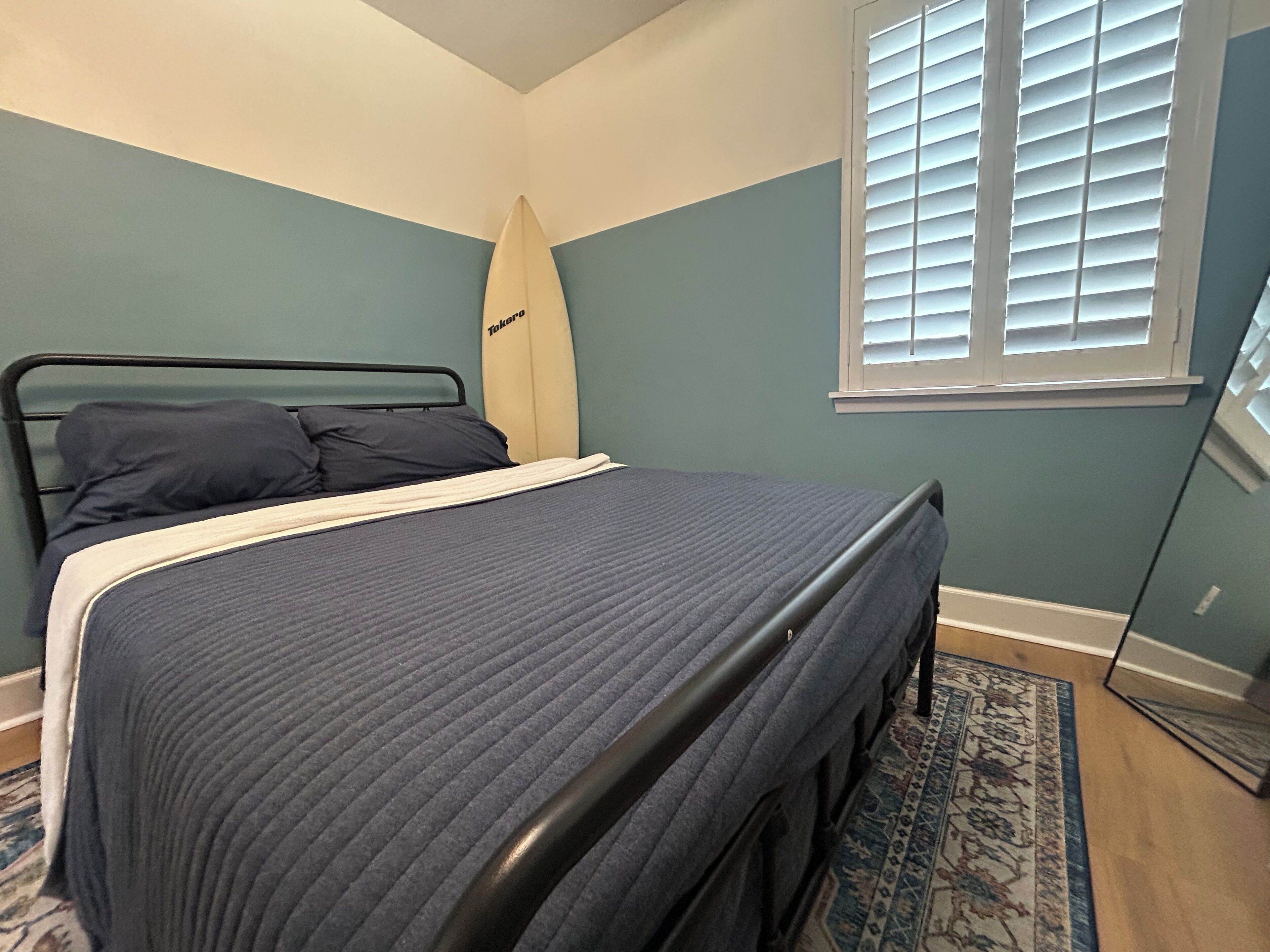 A bedroom with a bed, a surfboard, a window with blinds, and a patterned rug.