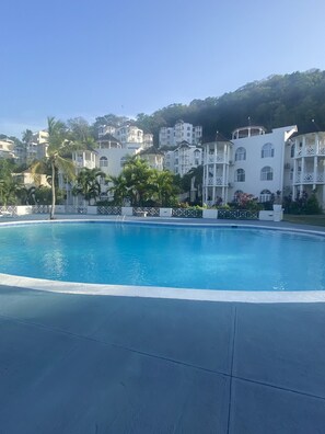 Ein Swimmingpool mit weißem Liegebereich, umgeben von mehrstöckigen Gebäuden mit Balkonen und einer hügeligen Landschaft im Hintergrund.