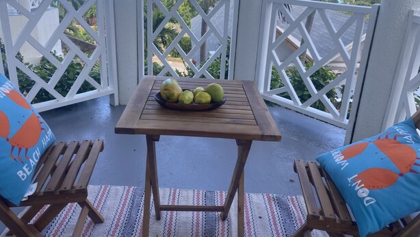 Een houten tafel met een fruitschaal op een balkon met witte lattenleuningen, twee houten stoelen met blauwe kussens en uitzicht op de oceaan.