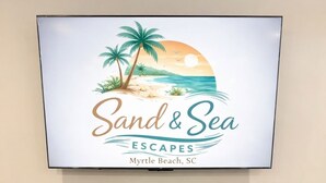 Un cuadro enmarco muestra una escena de playa con el texto 'Sand & Sea Escapes Myrtle Beach, SC' en una pared sobre un escritorio de madera blanca con tres cajones y un control remoto.