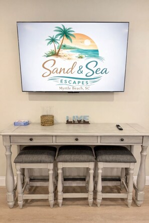 Ein gerahmtes Bild mit einer Strandansicht, darüber an der Wand der Text 'Sand & Sea Escapes Myrtle Beach, SC'. Darunter ein weißer Holztisch mit drei Schubladen und einer Fernbedienung.