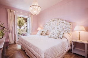 Ein Schlafzimmer mit einem großen Bett, einem Kronleuchter, einem Nachttisch und einem Fenster mit Vorhängen.
