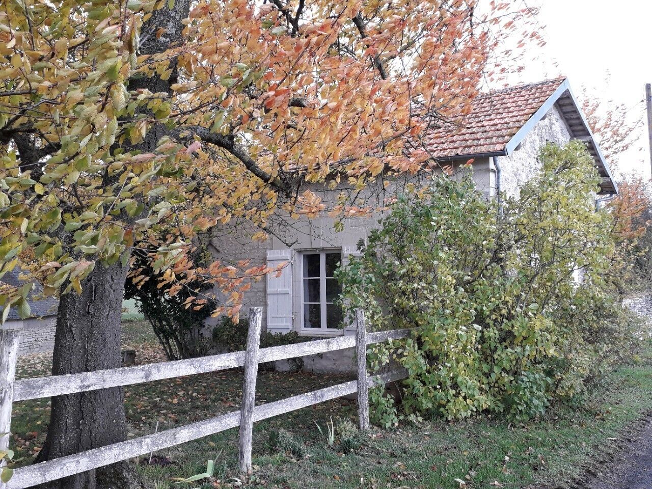 Un bâtiment rustique avec une façade en pierre, une clôture en bois et un arbre aux feuilles d’automne.