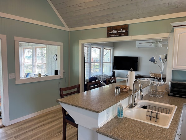 A kitchen with a granite countertop, a sink, a dining area with chairs, a television, and a sign above the counter reading 'macklin cabin EST. 2021'.