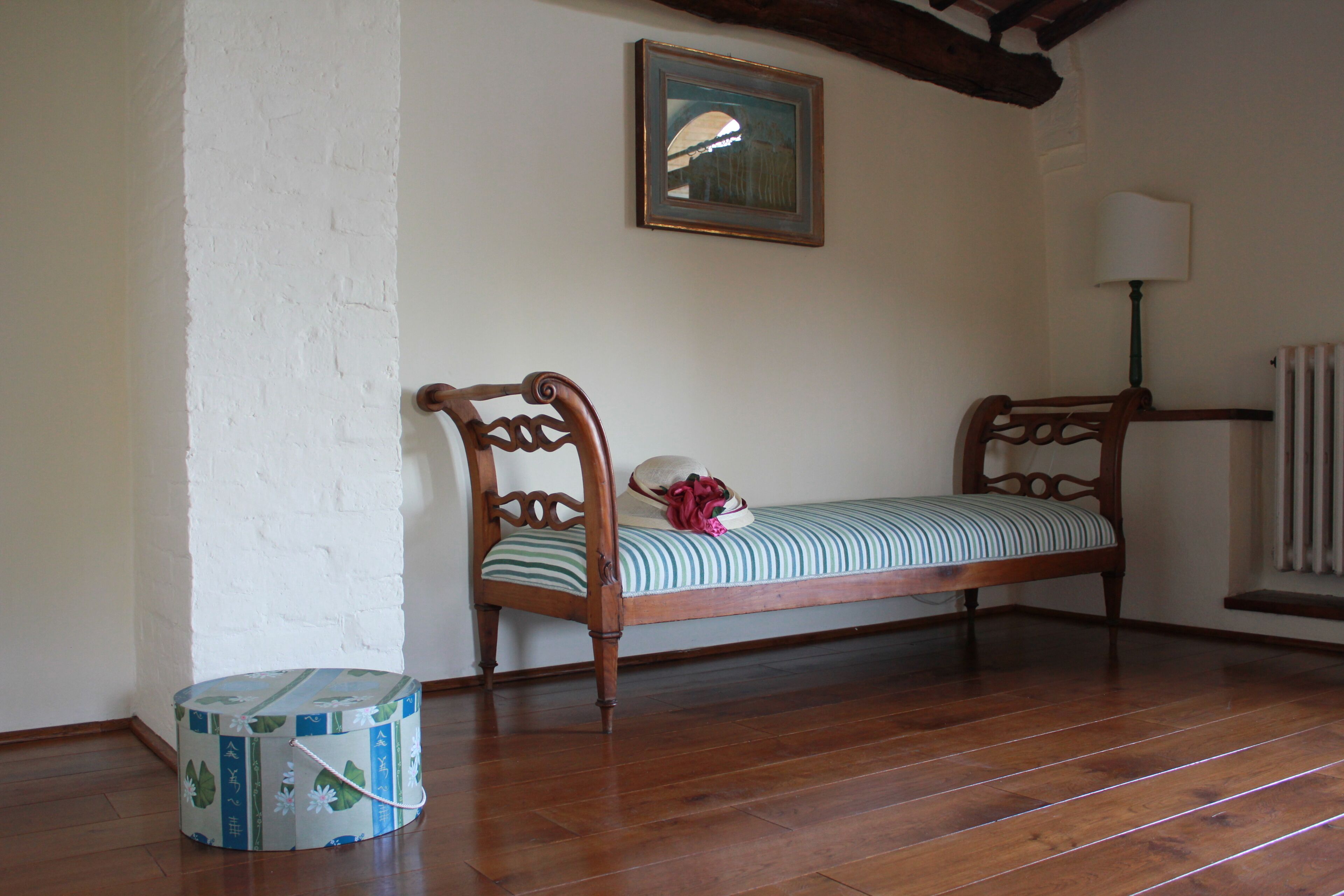 A room with a wooden bench, a round object on the floor, a framed picture on the wall, and a lamp.