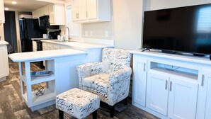 A modern kitchen with white cabinetry, a large TV, and a patterned ottoman.