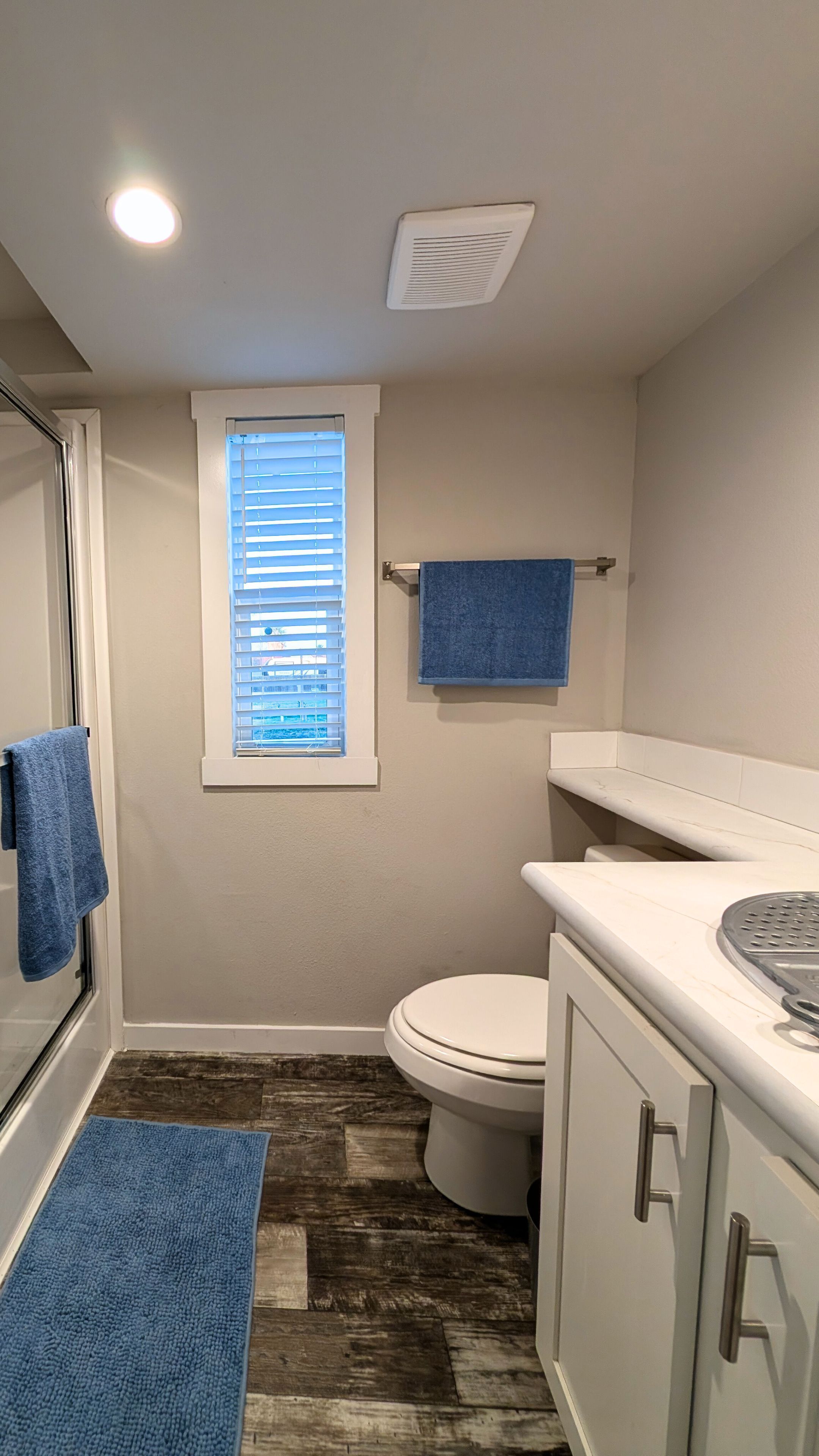 A bathroom with a toilet, a sink, a window with blinds, a towel rack, and a shower area.