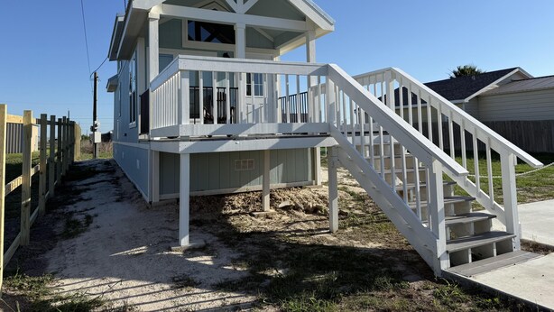 A beachfront house with a balcony, stairs, and a sandy ground.